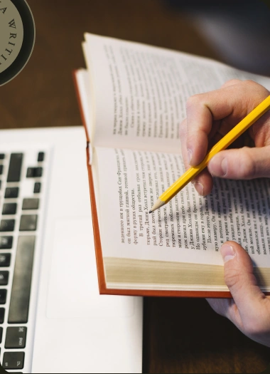 An editor reviewing a book with a pencil beside a laptop